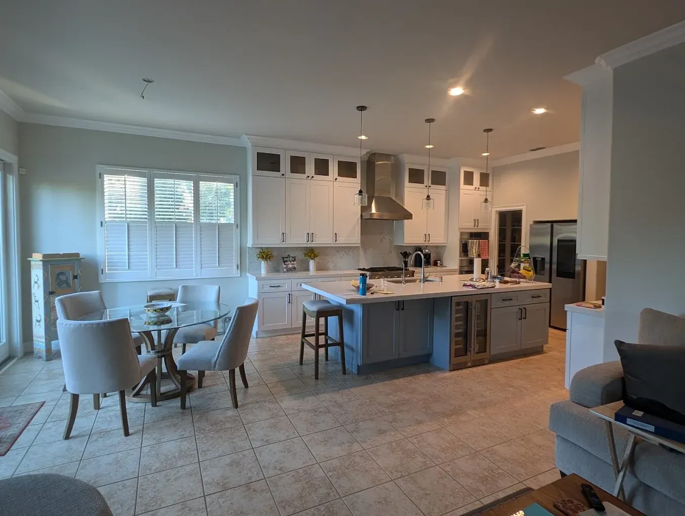 Spacious kitchen with island, white cabinets, and dining area. Light gray walls, window shutters, and tiled floor.