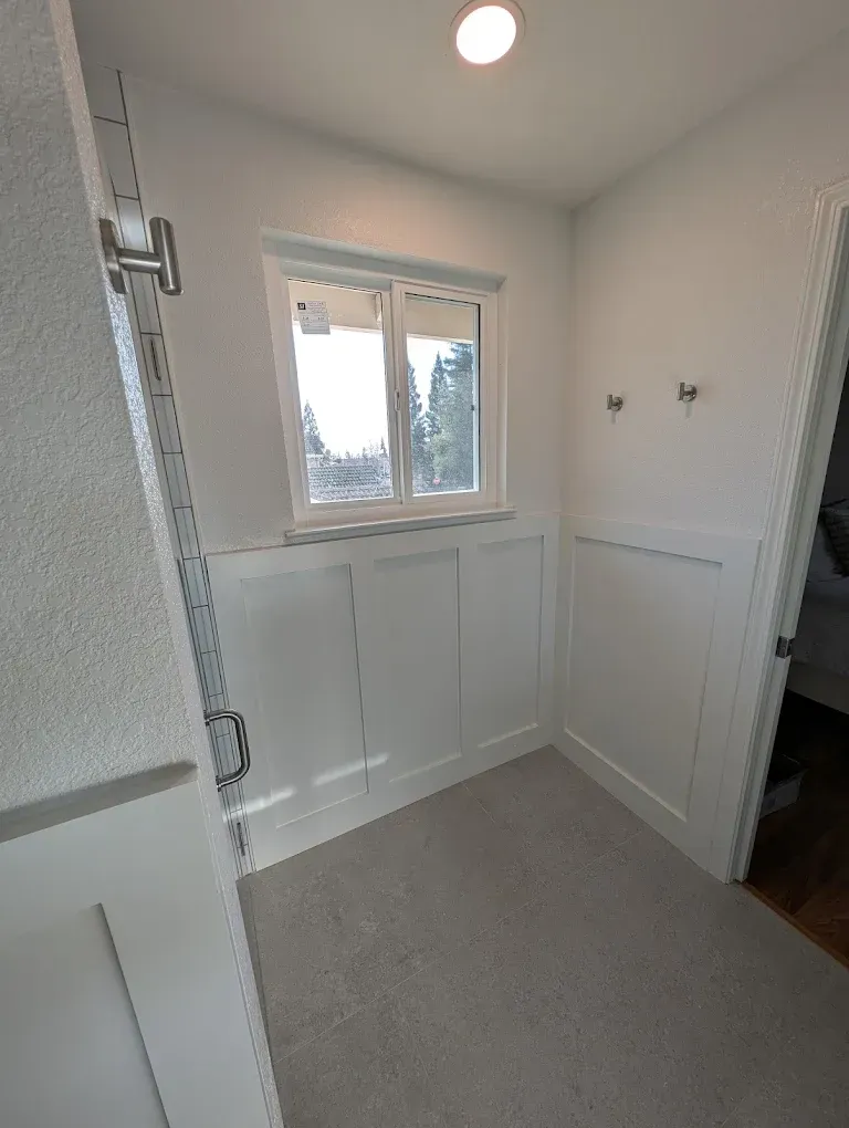 Small white bathroom with wainscoting, window, gray floor, and shower. Two hooks on the right wall.