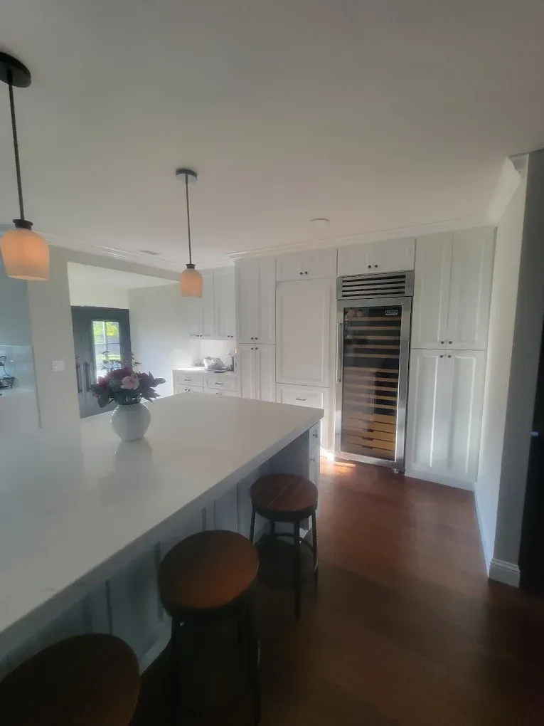 White kitchen with island, cabinets, and a wine fridge. Wooden bar stools sit near island.