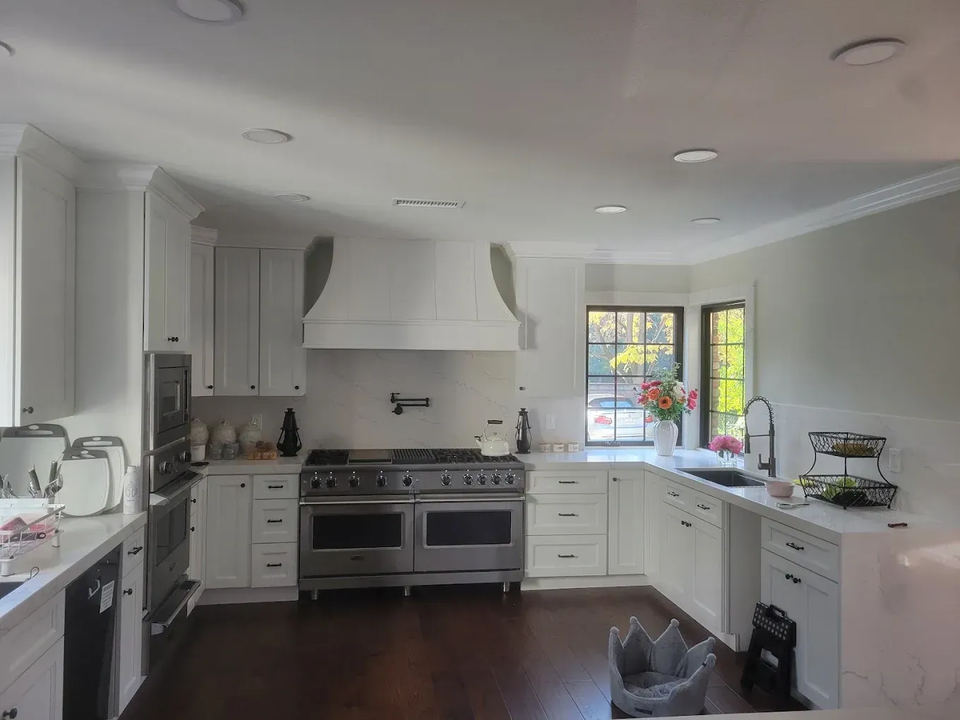 White kitchen with stainless steel appliances, dark wood floor, and a window with flowers.