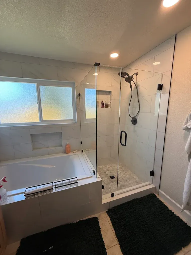 Modern bathroom with glass shower, bathtub, and light-colored tile. Dark green rugs on floor, black shower hardware.