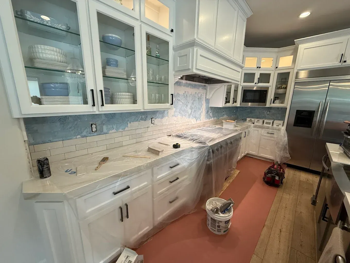 Kitchen under renovation: white cabinets, blue backsplash being installed, appliances covered in plastic.