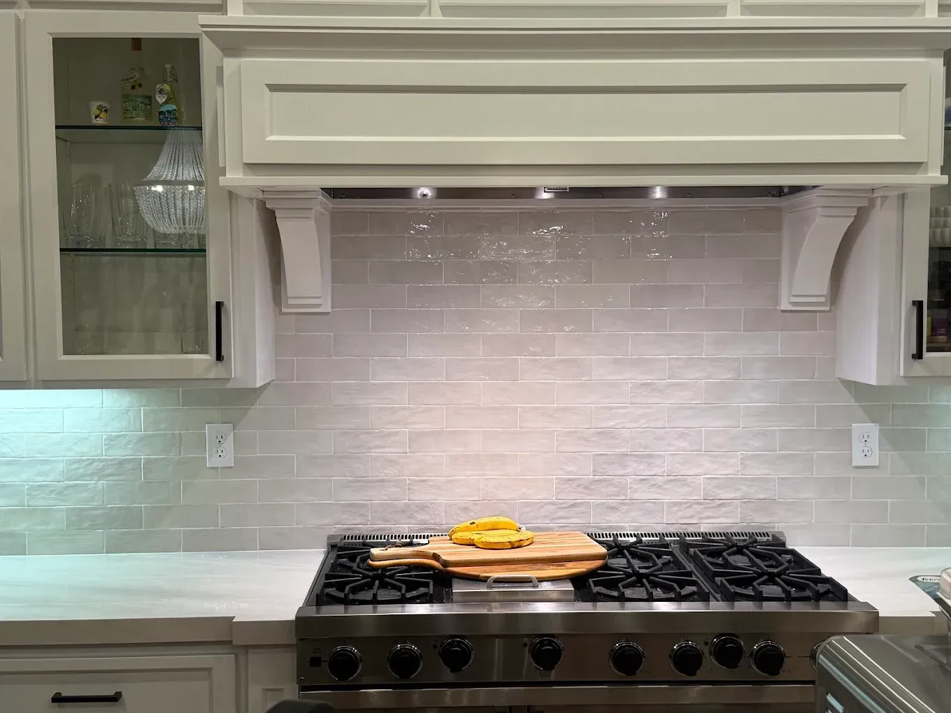 Kitchen with stove, light gray backsplash, and light wood cutting board with food on it.
