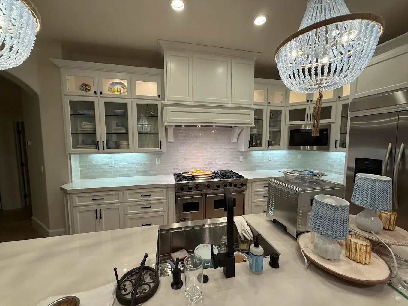 White kitchen with glass-front cabinets, range, and two crystal chandeliers.