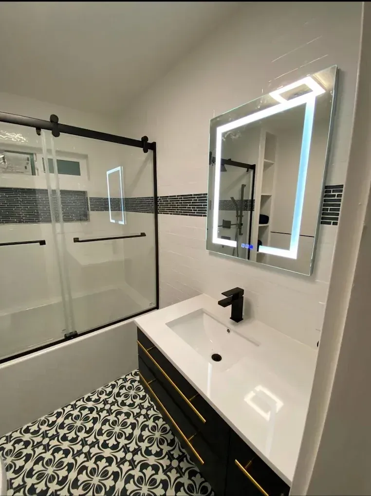 Modern bathroom with black and gold vanity, lit mirror, tiled floor, and glass shower.