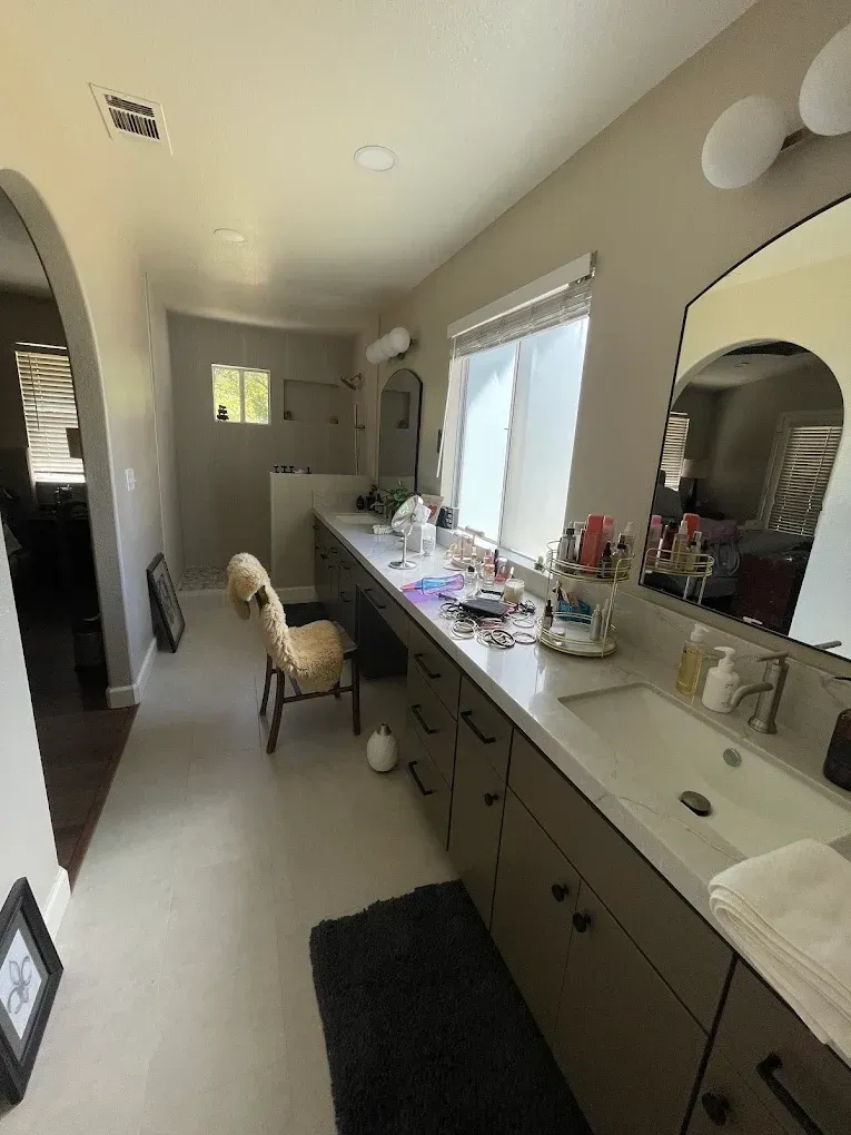 Long, light-toned bathroom with a vanity and two sinks; mirror, chair, and window.