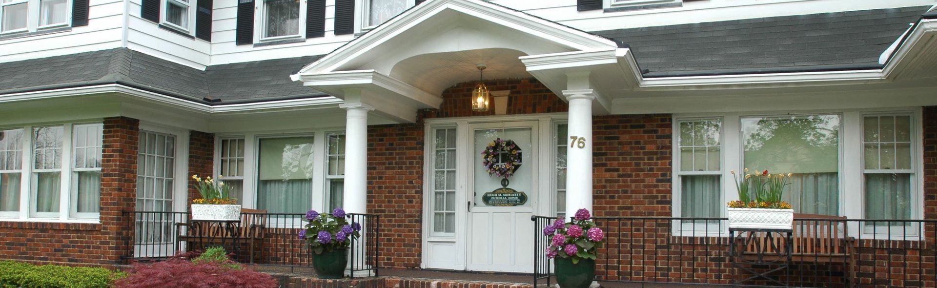 Front entrance of a white house with green shutters, brick steps, and flower pots by the doorway