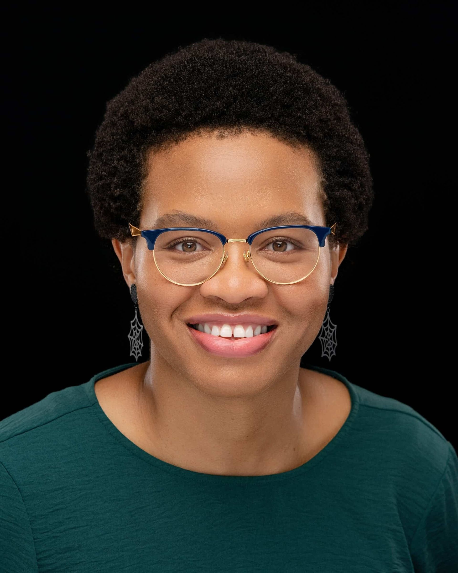 Professional headshot of Melinda Estrada wearing a dark green crew-neck top with geometric dangling earrings. The individual has short, tightly curled hair. The background is solid black, creating a professional studio portrait setting.