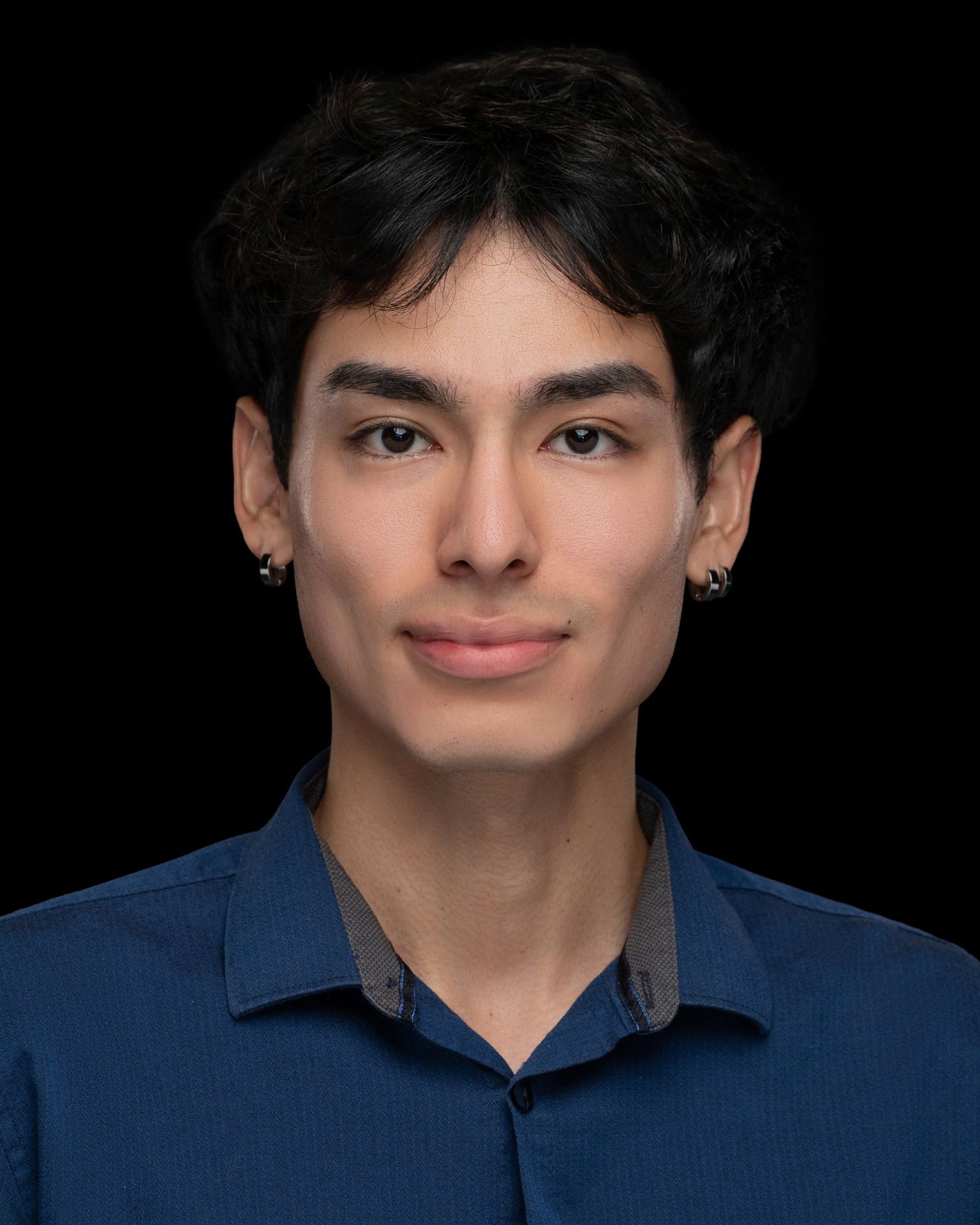 Portrait of a person with dark hair wearing a blue collared shirt and earrings, posed against a black background.