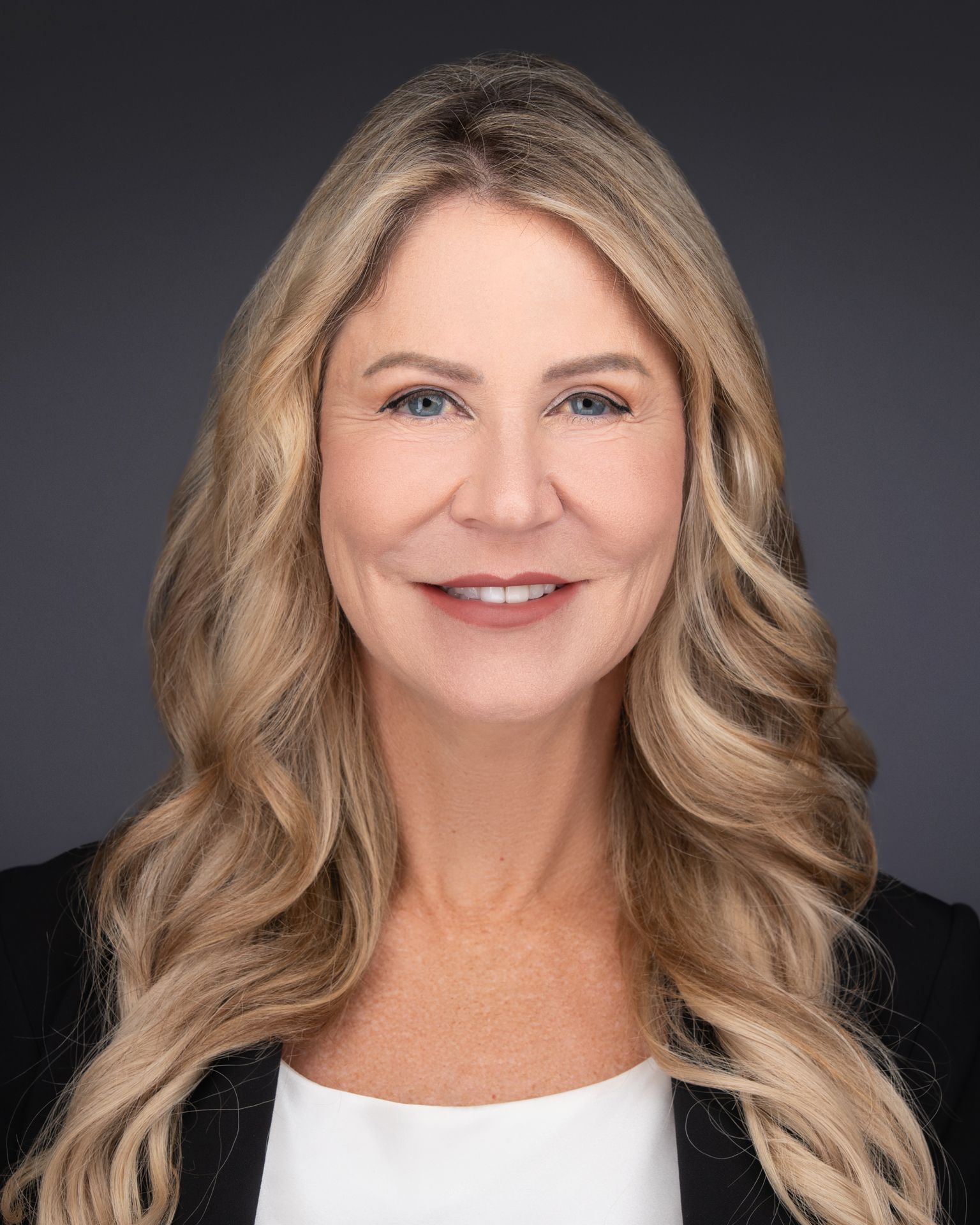 Professional Headshot of Rylee Hickman with long, voluminous curly hair wearing a black top layered over a patterned high-neck shirt in shades of blue and gray. A delicate gold necklace with a Y-shaped pendant is visible. The background is solid black, creating a professional studio portrait setting.