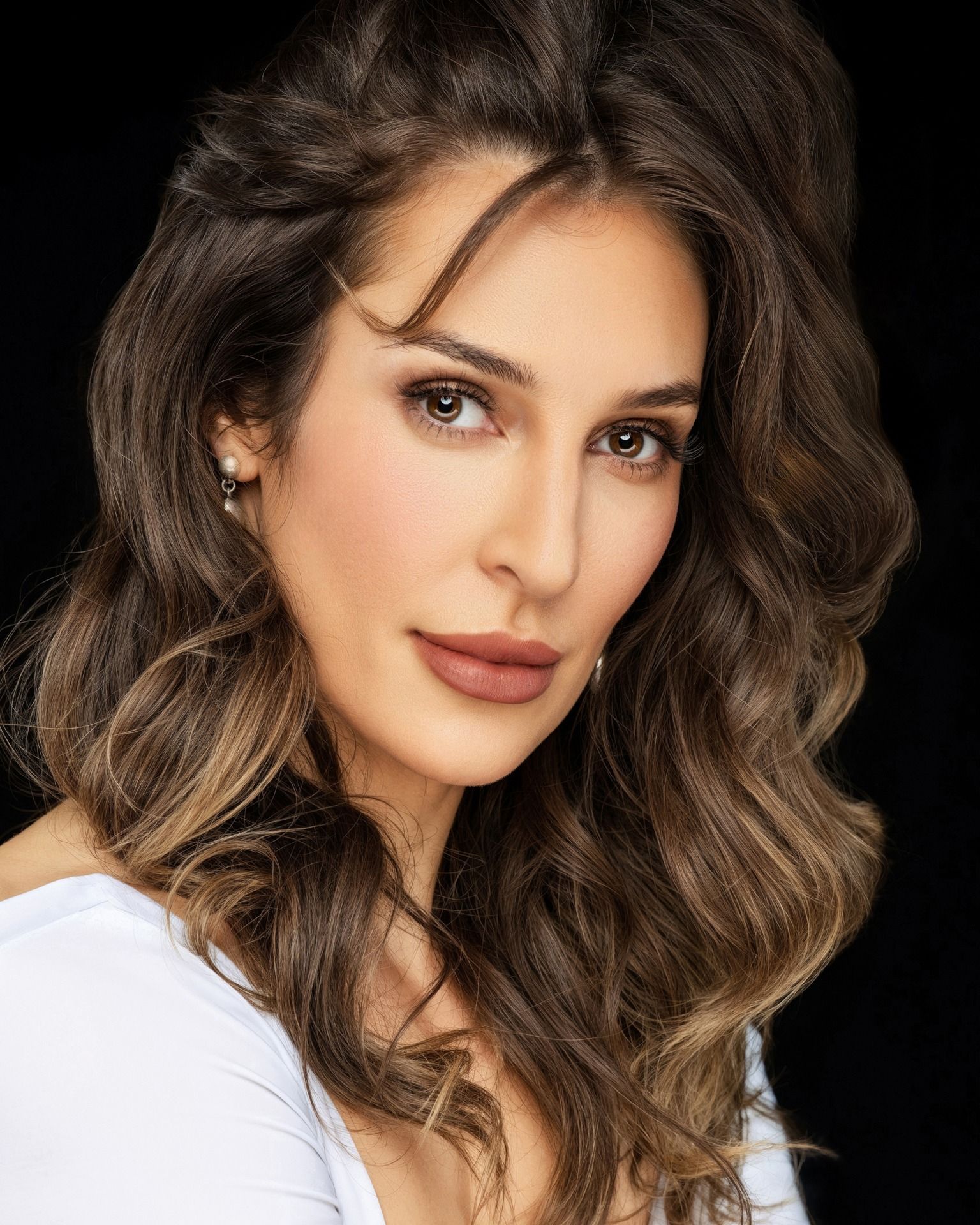 Portrait of a person with wavy brown hair wearing a white top, looking directly at the camera against a black background.