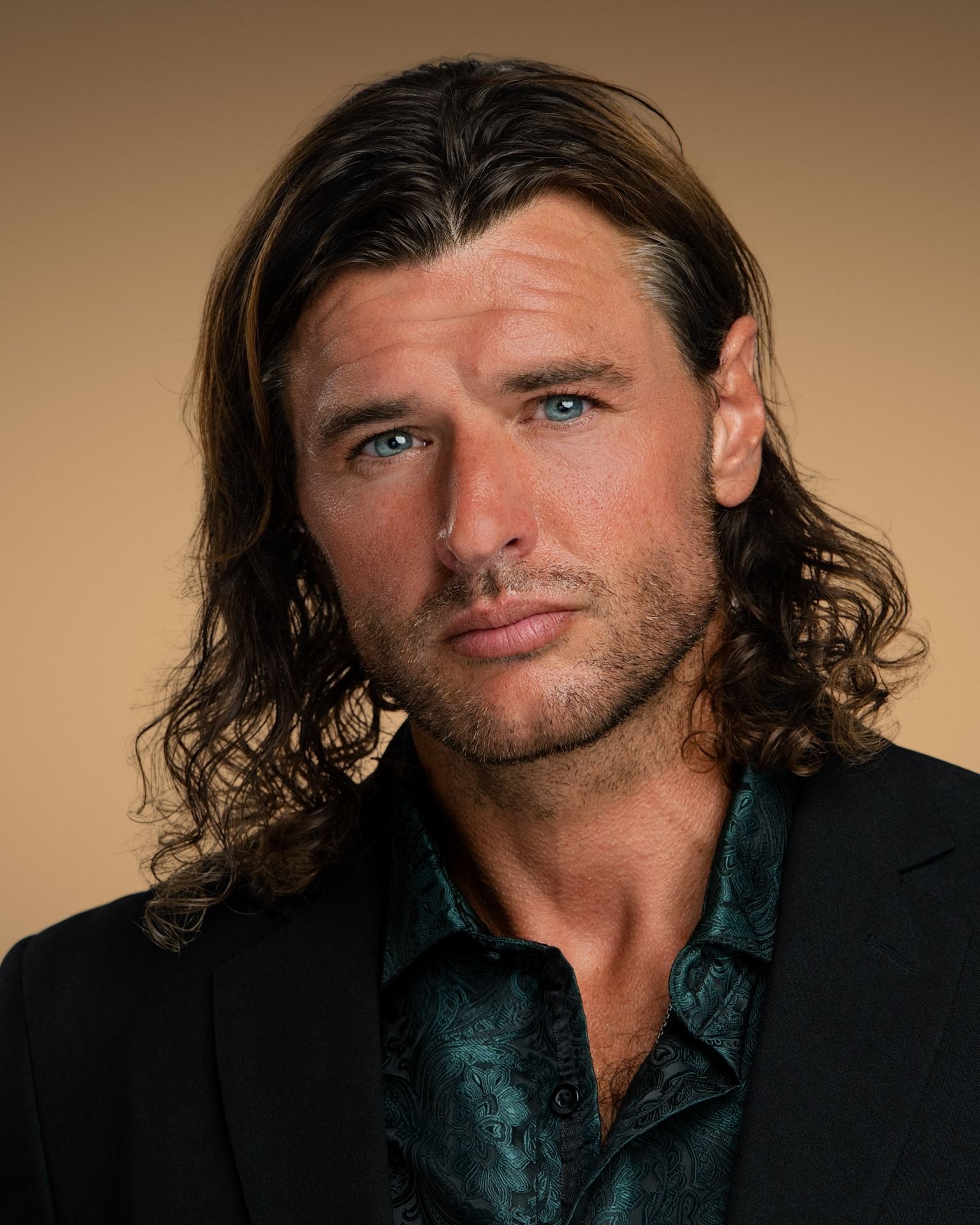 Portrait of a person with long wavy hair wearing a dark blazer and patterned shirt, posed against a beige background.