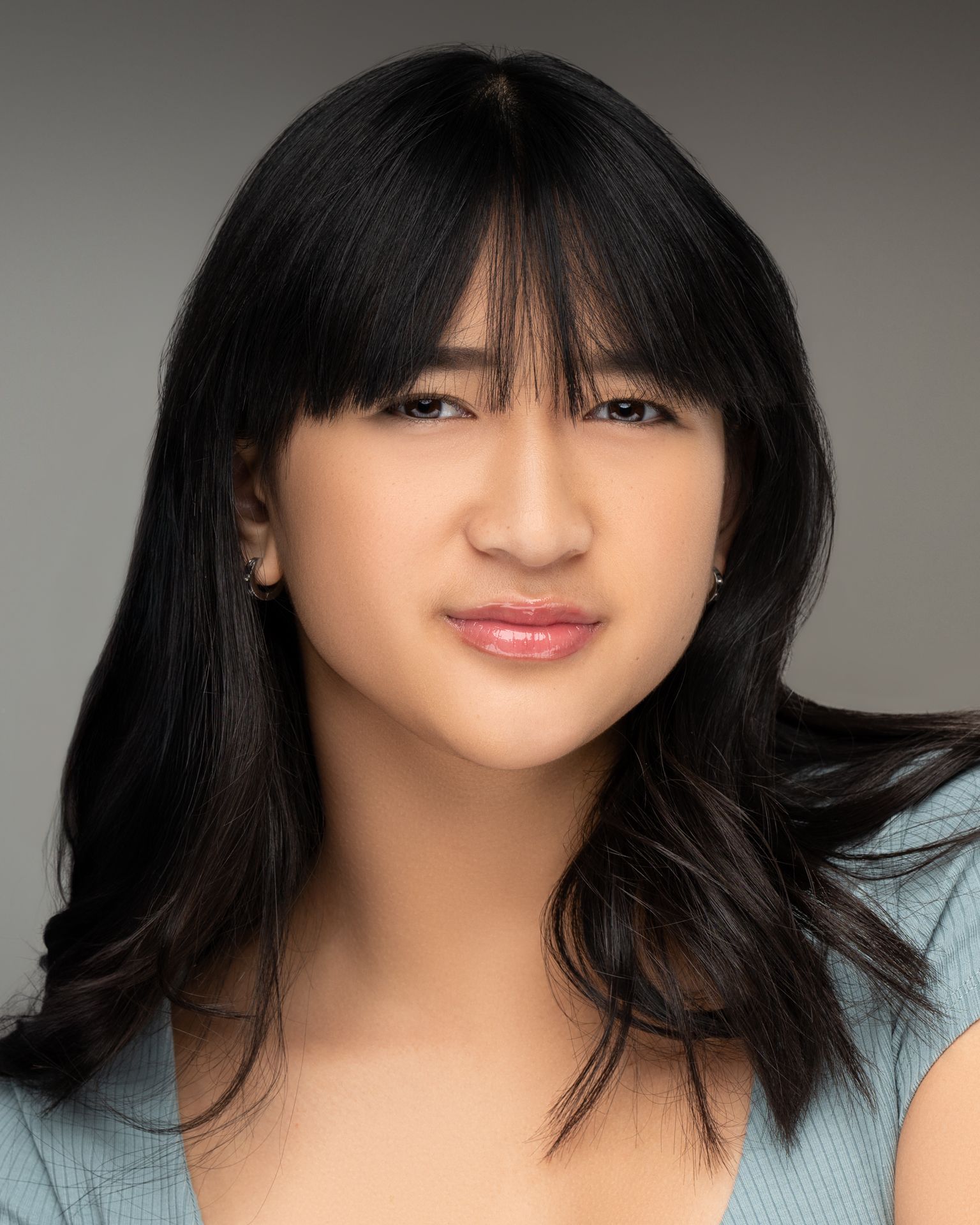 Professional headshot of a person with long dark hair in a light blue top against a gray background.