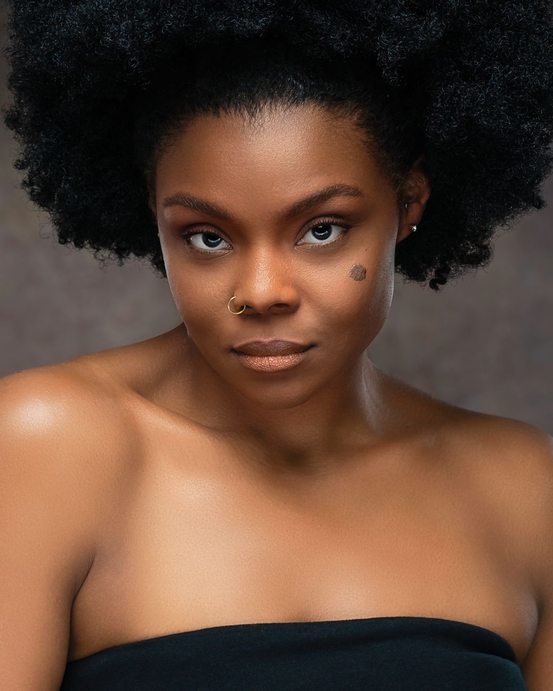 Portrait of a person with an afro hairstyle, nose ring, and strapless top, looking directly at the camera against a neutral background.