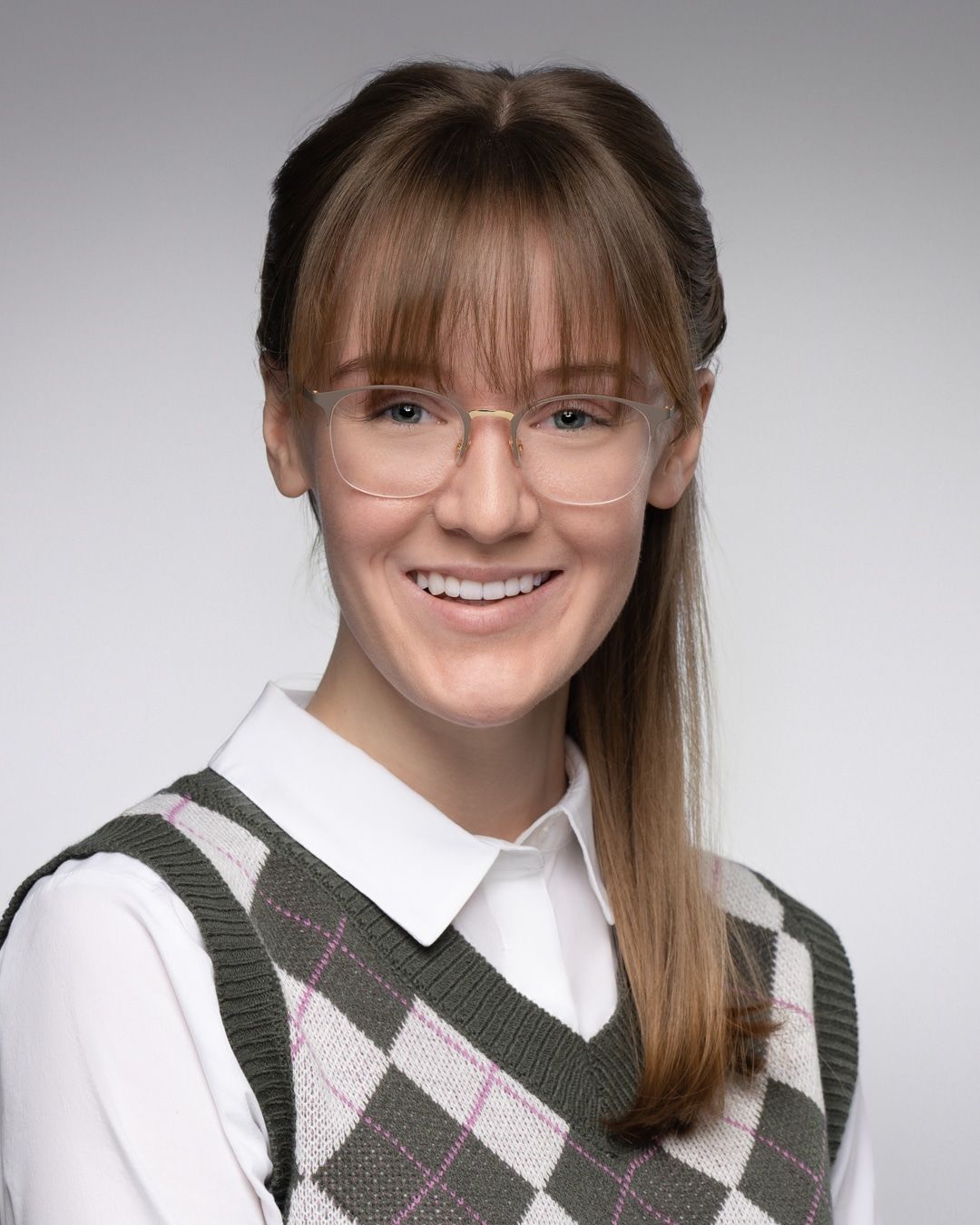 Person wearing glasses, a white shirt, and an argyle sweater vest, smiling against a gray background.