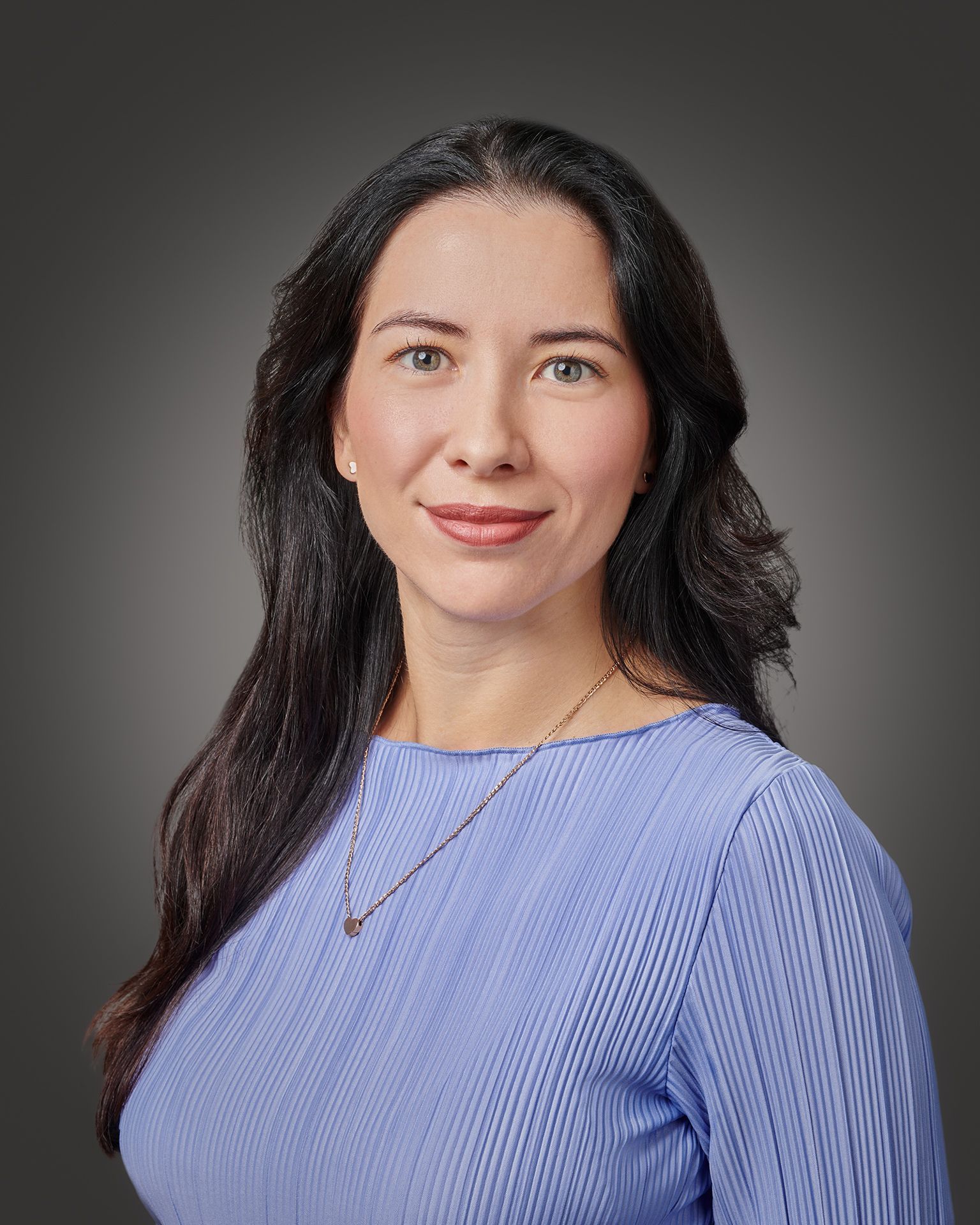 Professional Headshot of Lauren Mendez with shoulder-length, layered brown hair wearing a black blazer over a light gray top. A delicate gold necklace with a rectangular pendant is visible. The background is solid black, creating a professional studio portrait setting.
