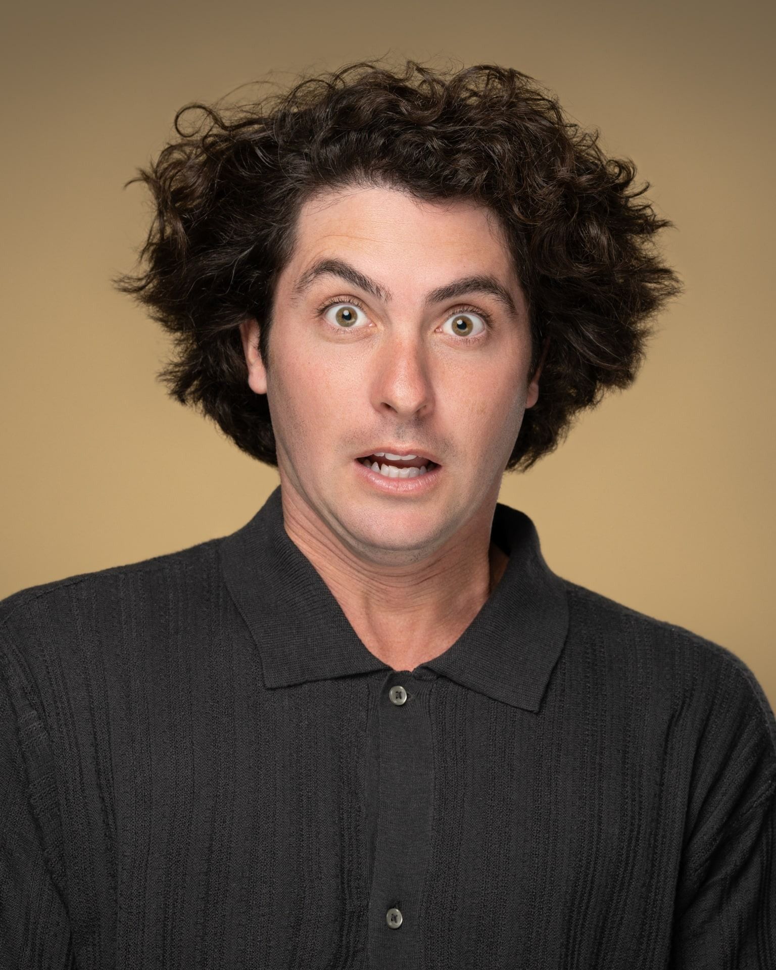 Professional headshot with curly hair wearing a blue and black plaid shirt on a dark studio background.