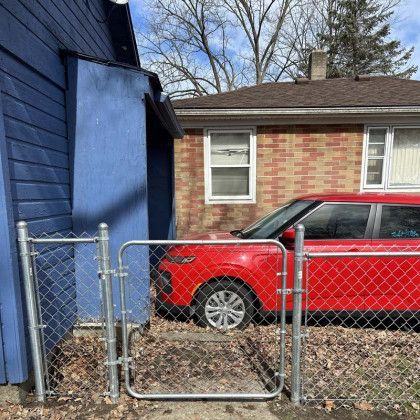 Red car parked in front of a brick house. Chain-link fence with open gate near a blue building.
