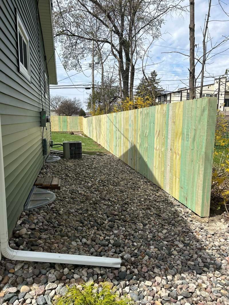 Green house siding next to a light green fence made of wooden planks, a bed of rocks, and a white gutter.