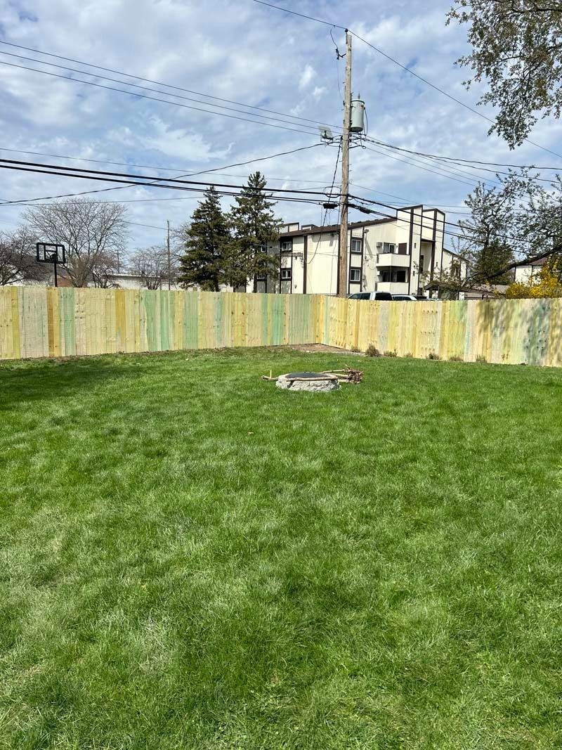 Green backyard with fire pit and colorful fence; buildings and sky in the background.