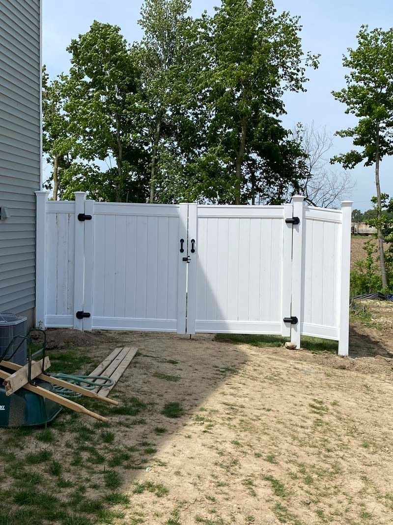 White vinyl fence with double gate and black hardware, in a yard with trees.