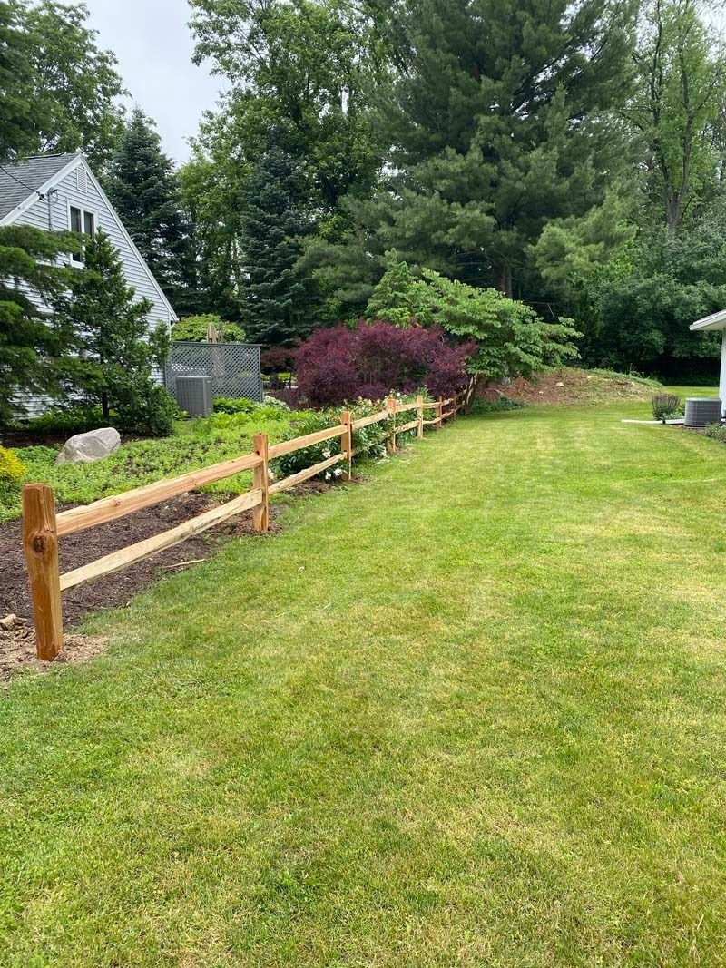 Wooden split-rail fence in a grassy yard, with trees and a house in the background.
