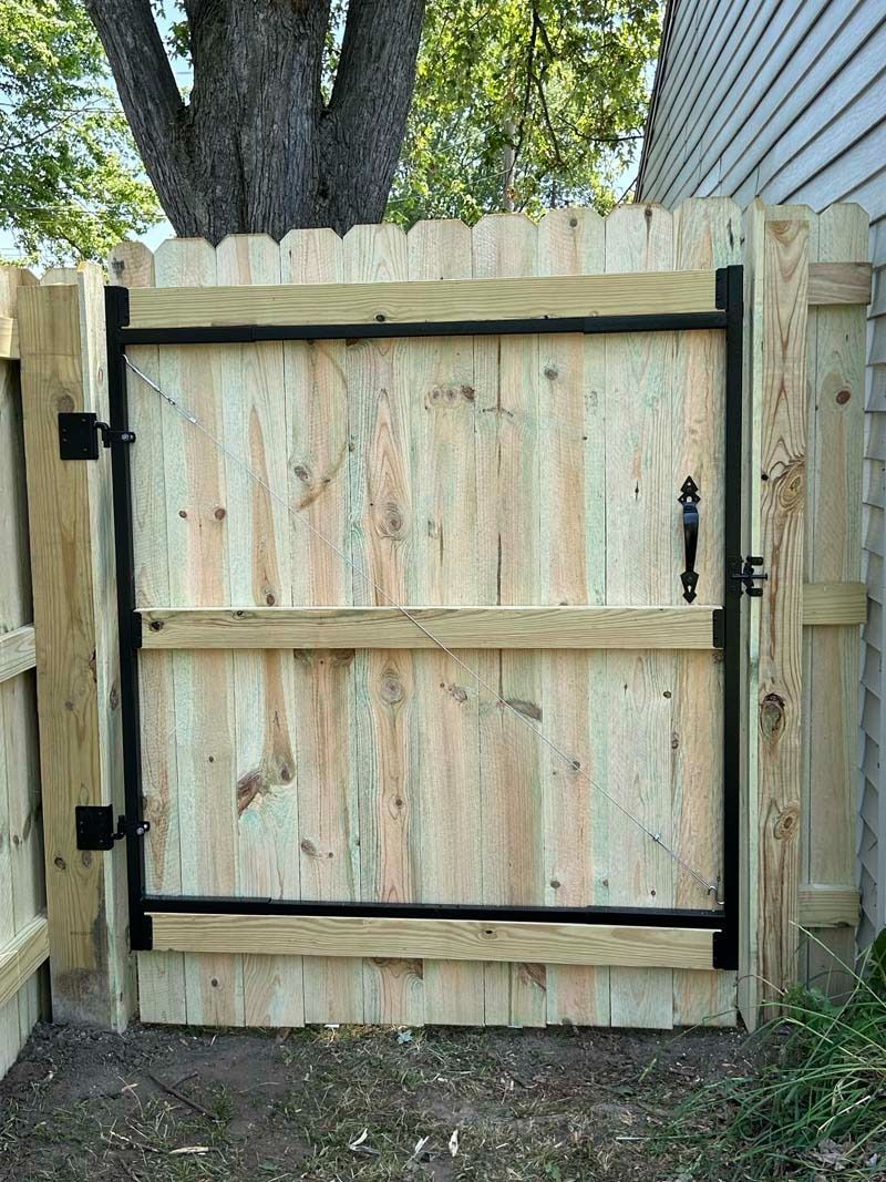 Wooden gate in a wooden fence, with a black metal frame, handle, and hinges.