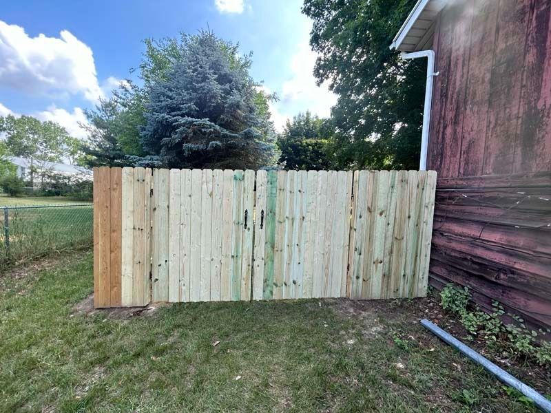A wooden fence, partially constructed, sits next to a red building on a grassy lawn.