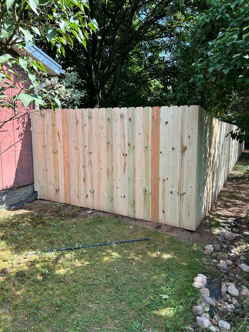 New wooden fence in a grassy yard, against a red building, trees in the background.