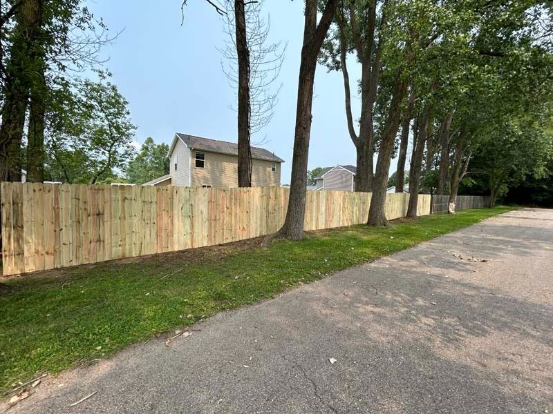 A new wooden fence lines a road, separating a grassy yard from the pavement, trees, and a house in the background.