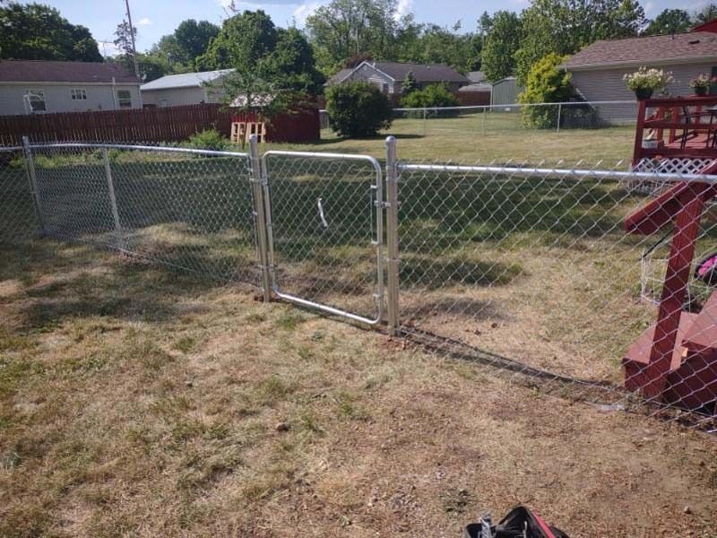 A chain-link fence and gate in a yard, with a red wooden deck and houses in the background.
