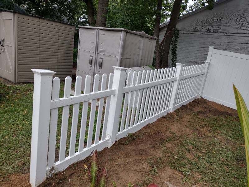 White picket fence in a yard, with sheds and a tree in the background.