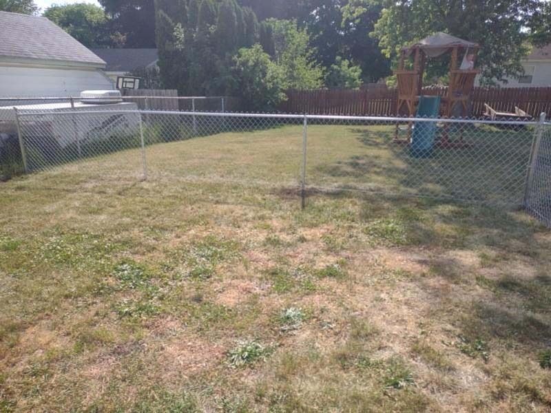 Chain-link fenced backyard with dry grass, a playset, and parts of homes.