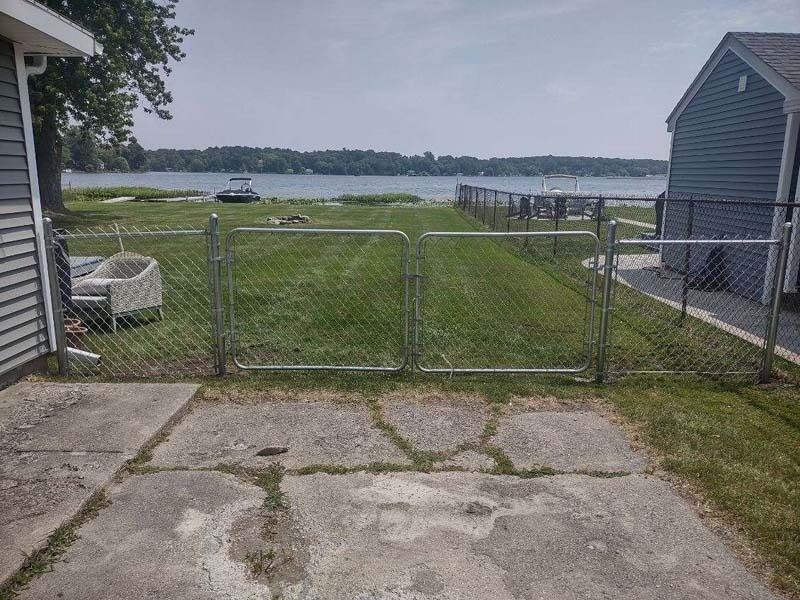 Chain-link fence separates a grassy yard from a lake, between two houses.