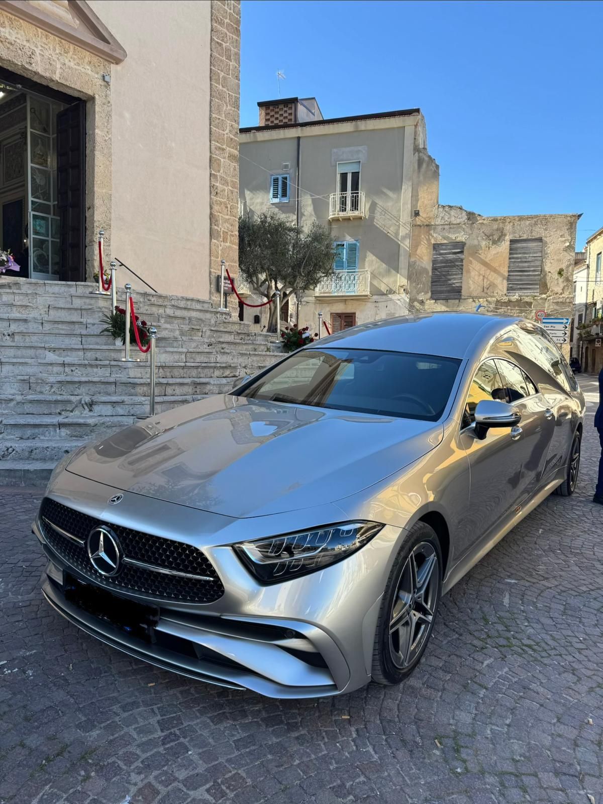 Una berlina Mercedes color argento parcheggiata su una stretta strada lastricata, accanto a vecchi edifici e scale.