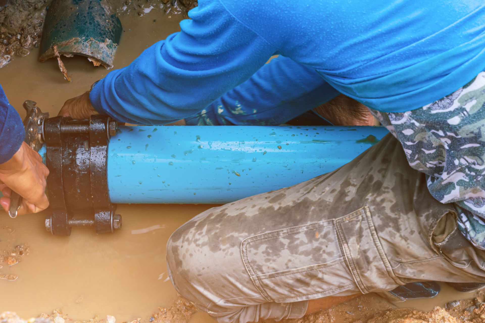 Technician repairing a damaged underground water line in a muddy trench using pipe fittings