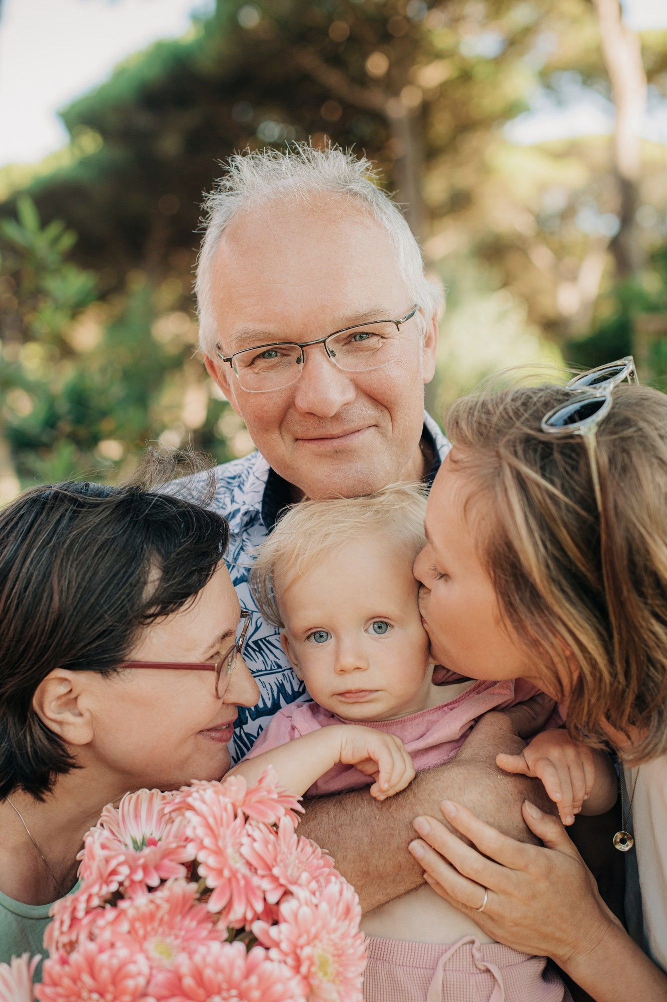 Family portrait: Man with glasses and two women kissing a toddler holding pink flowers, outdoors.