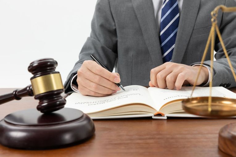 Gavel, scales of justice, and a person in a suit writing in a book on a wooden desk.