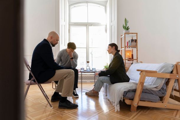 Two people in therapy session; one client holds head; therapist listens. Light-filled room with window, sofa.