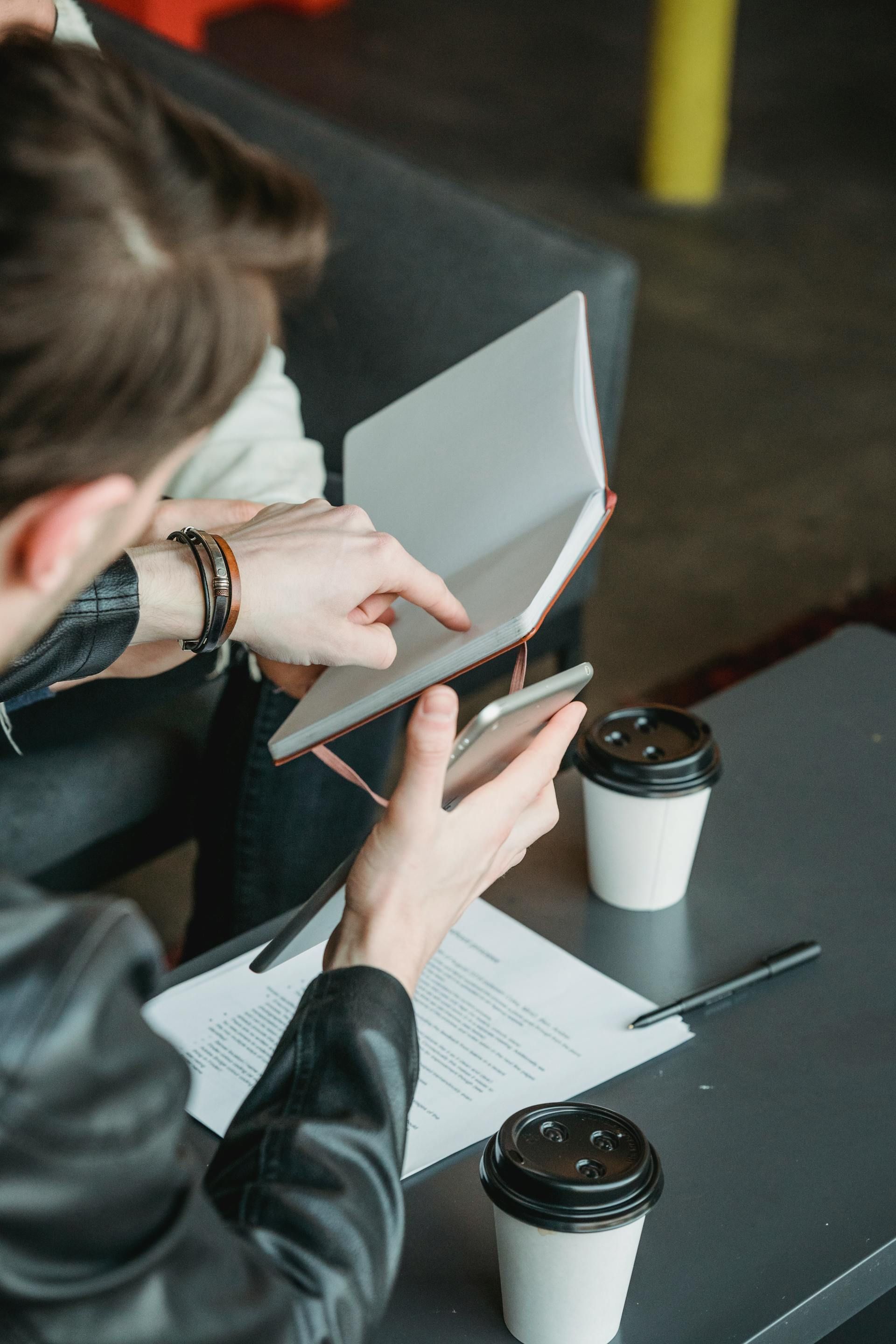 Two people looking at a notebook and phone at a table with coffee cups and papers.