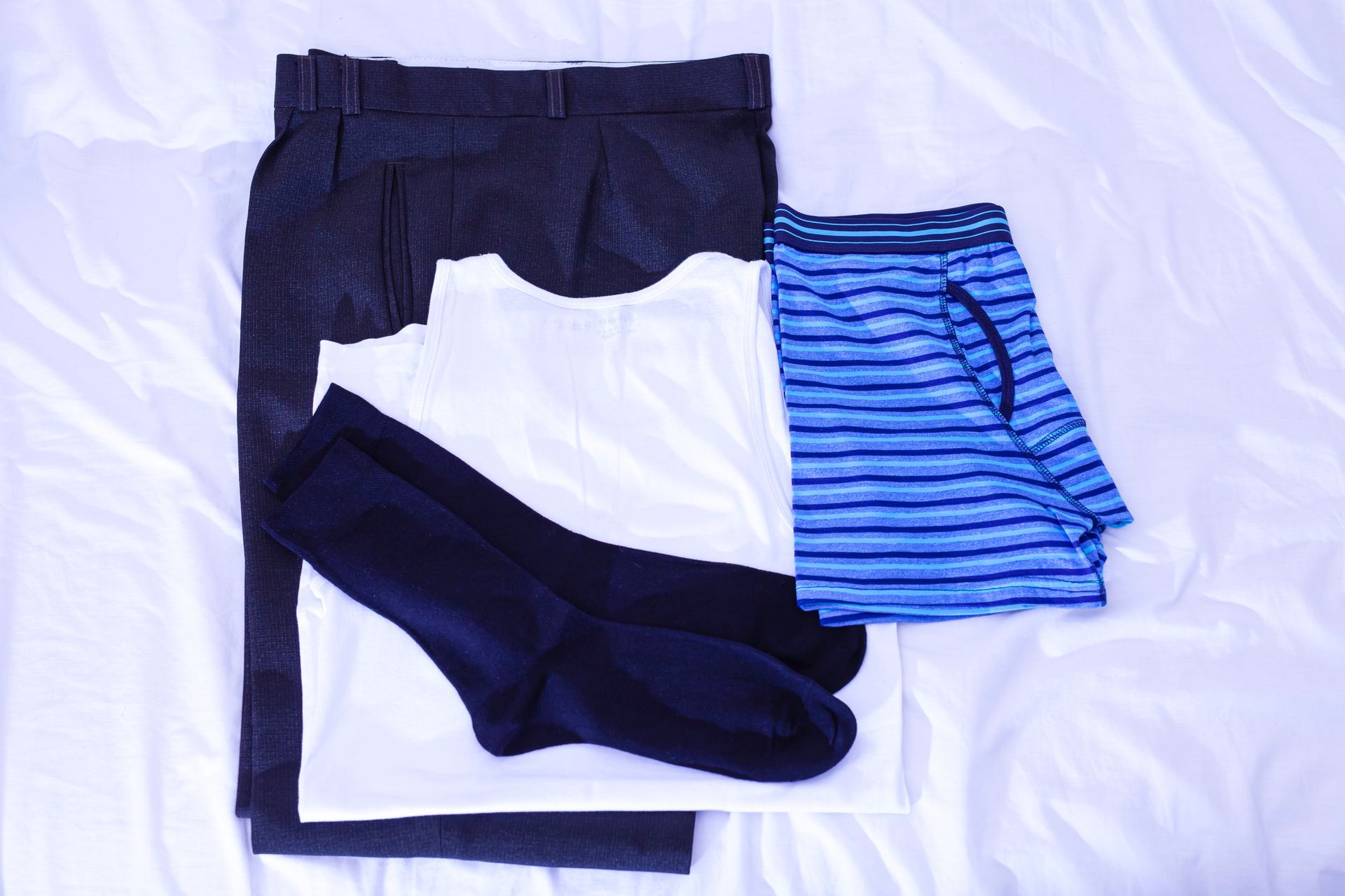 Vestiti stesi sul letto: pantaloni scuri, camicia bianca, biancheria intima a righe blu, calzini scuri.