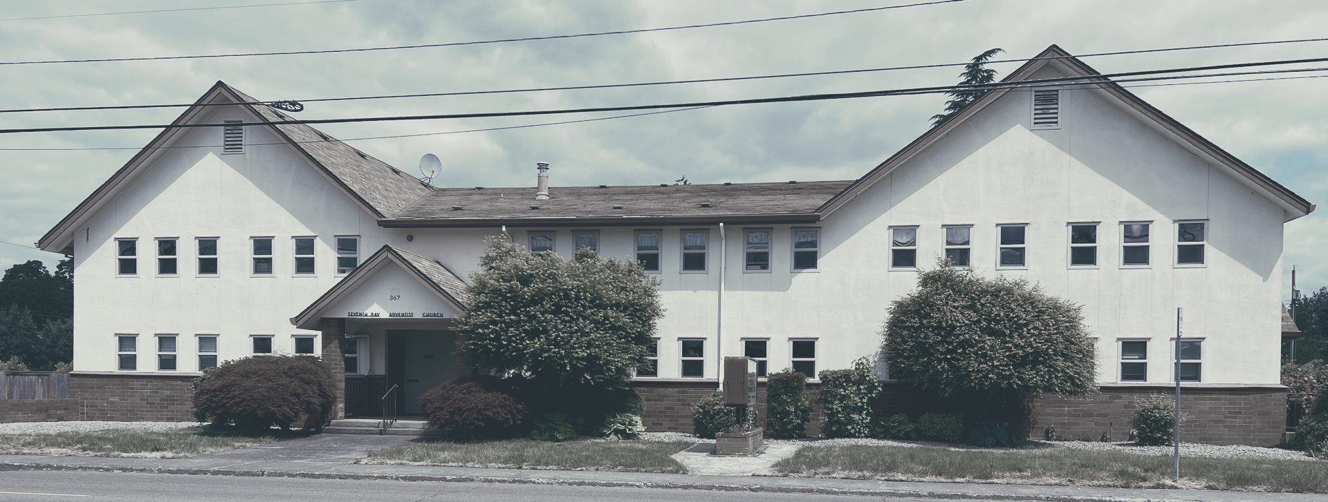 White two-story building with multiple windows, bushes in front, and a cloudy sky.