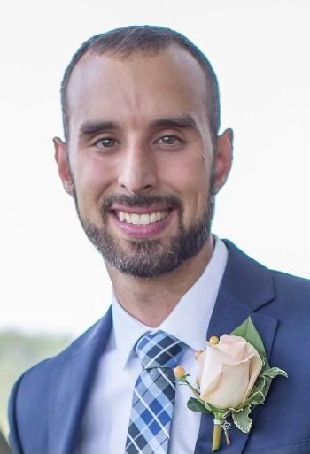 Man in a blue suit with a blue and white checkered tie smiling, wearing a rose boutonniere.