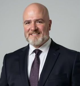 Bald man with a graying beard, wearing a dark suit and tie, smiling at the camera.