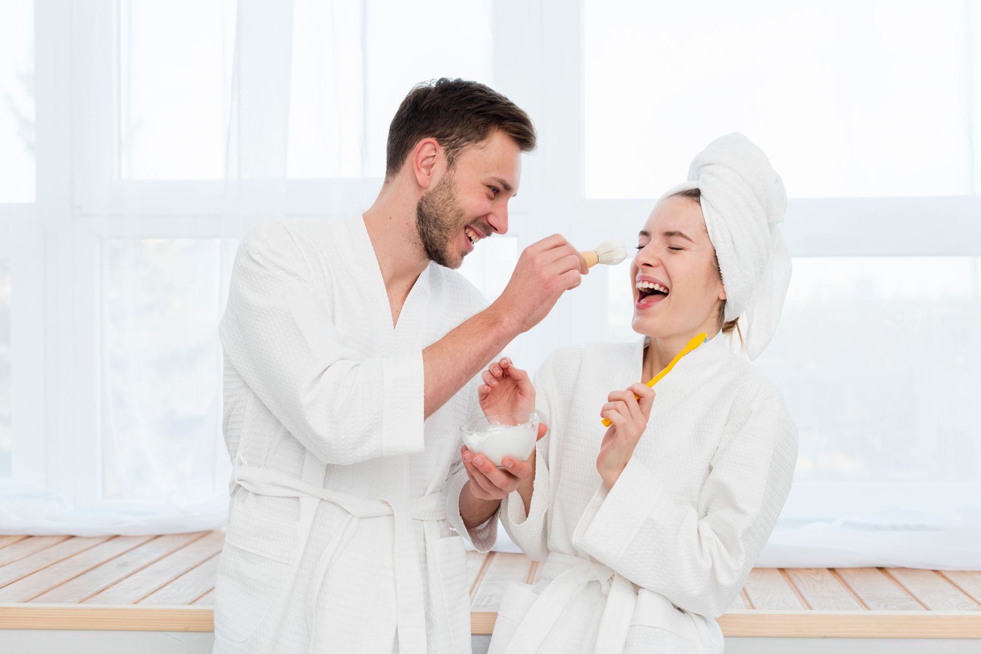 Two people in white bathrobes share ice cream and laugh in a bright spa setting