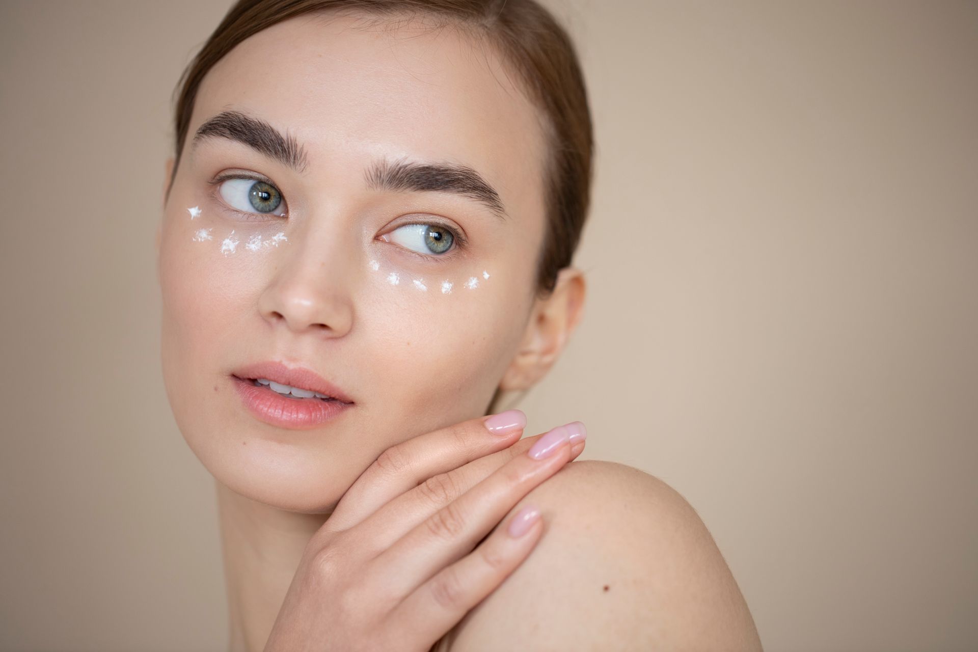 Woman with eye cream applied under eyes, looking to the side, touching her shoulder.