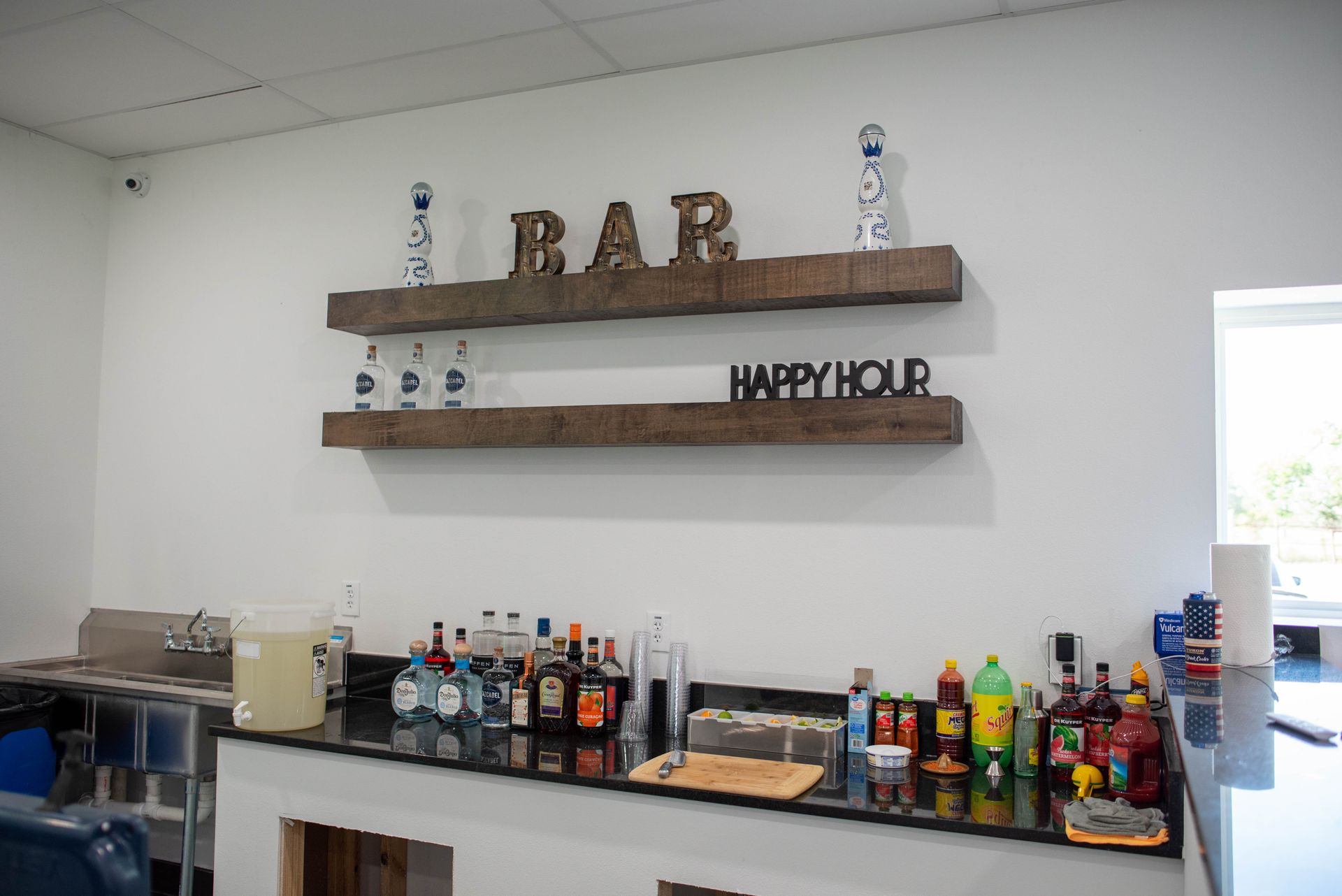 Bar counter with liquor bottles and a wooden shelf sign reading “BAR” and “HAPPY HOUR” in a white room