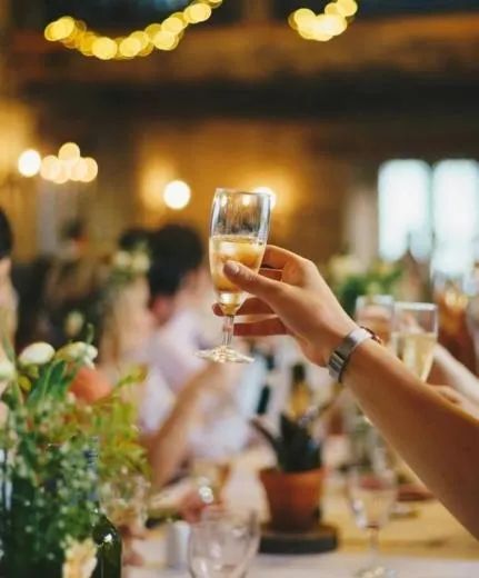 Hand holding a champagne glass at a warmly lit dinner party with blurred guests and floral decor