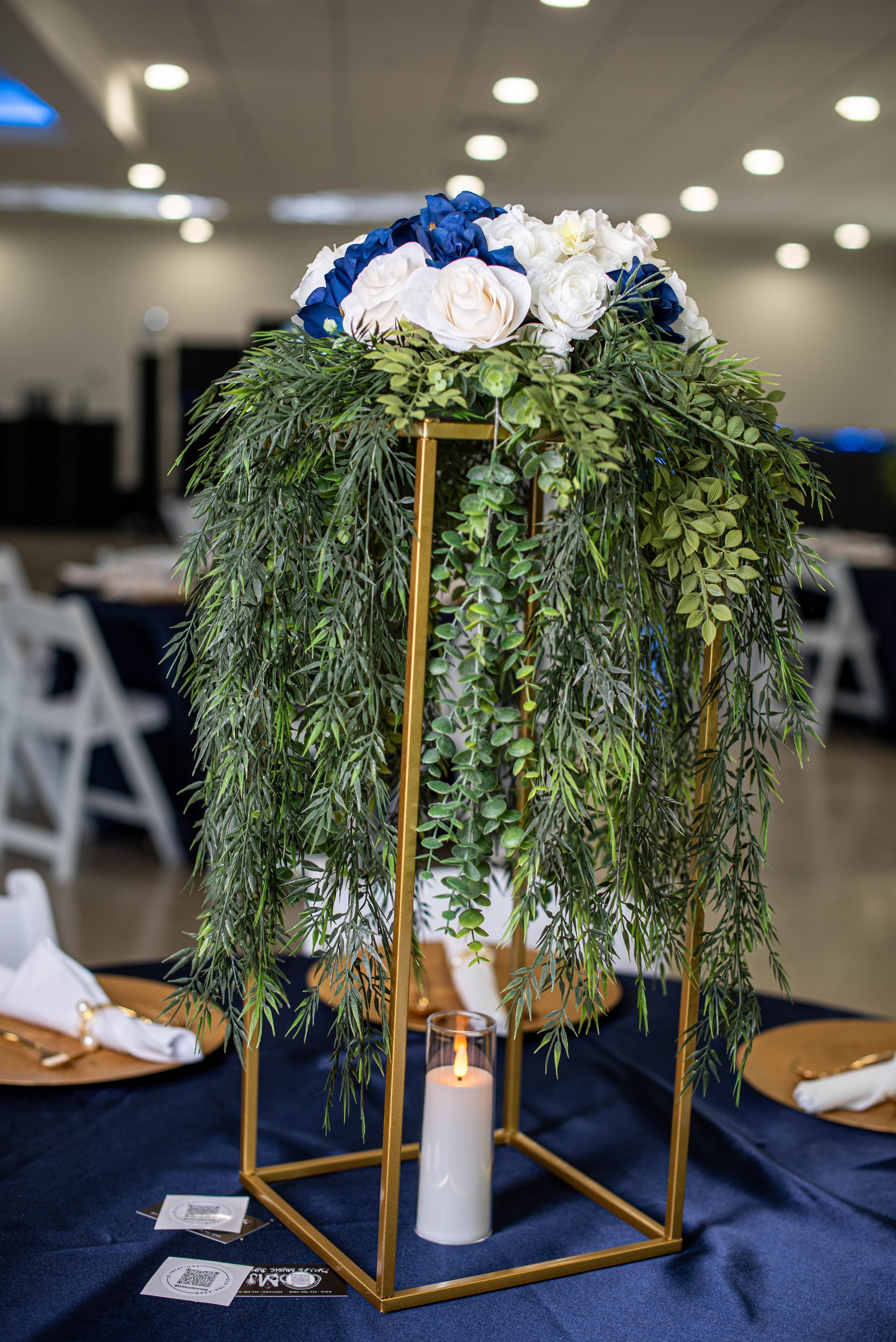 Gold candle stand with cascading greenery and white flowers on a navy tablecloth, with a lit candle below