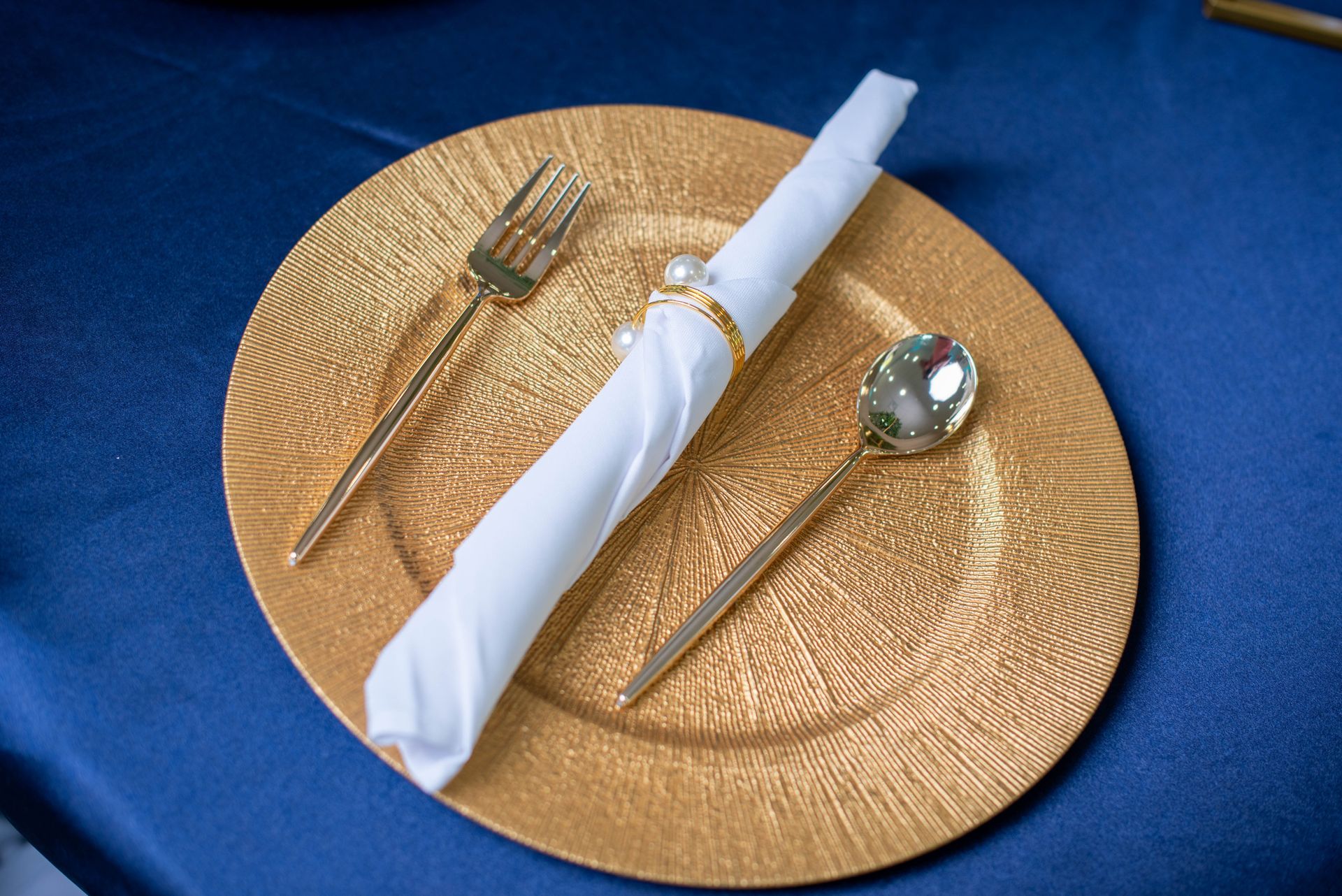 Gold charger plate with rolled white napkin, fork, and spoon on a blue tablecloth